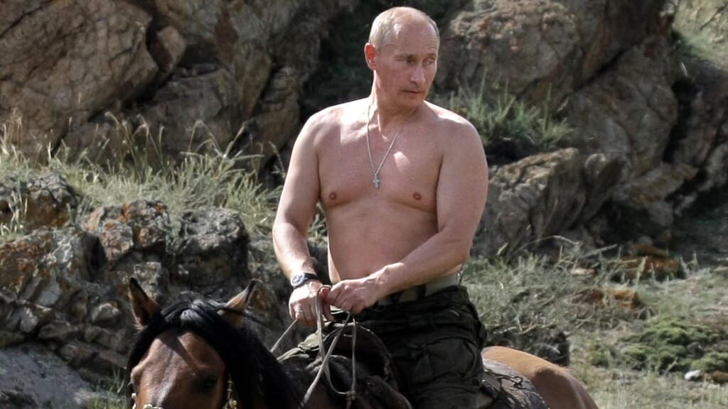Russian autocrat Vladimir Putin holds the reins of a brown horse as he rides bare-chested through a low-scrub landscape in 2012. [Source The Daily Beast]
