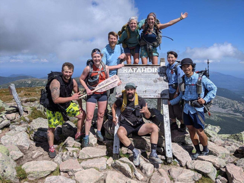 Brendan (far left) celebrates with his Appalachian Trail family on the summit of Mt. Katahdin, the AT’s northern terminus in Maine. Eight men and women wearing boots, shorts, bandanas, and assorted hiker garb pose around the summit sign post. It’s a clear summer day on Maine’s highest mountain.