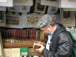 Mark Willis peruses a 1745 volume by Voltaire at a bouquiniste book stall on the banks of the Seine in Paris. He wears a brown leather jacket and checkered flat cap. He holds the open book in his hands. Rows of old books are seen on shelves behind him. [2005 photo by Ms. Modigliani]