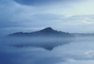 Fog at Isle Royale. The largest island in Lake Superior, obscures the edges of land, water, and sky. The photo is a mix of blue and gray tones. Fog in the foreground makes water and sky into one blurred element. An outer island in the distance appears as an indistinct land mass. The photo’s overall effect resembles the author’s waning peripheral vision, which he often compares to impressionist paintings by Claude Monet.