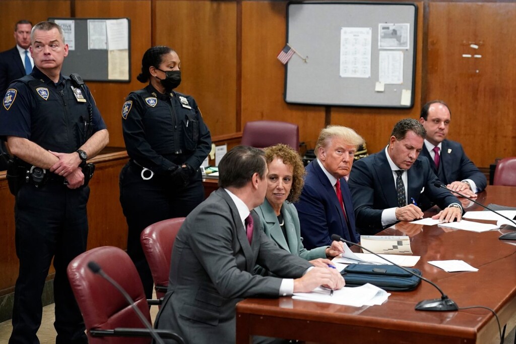 Donald Trump sits at the defendant’s table at his criminal arraignment in Manhattan District Court on April 4, 2023. He wears a dark suit with red tie. Flanking him are several defense attorneys wearing lawyerly professional attire. Behind him stand two stalwart-looking New York Police officers in dark blue uniforms. One wears a COVID-19 face mask.